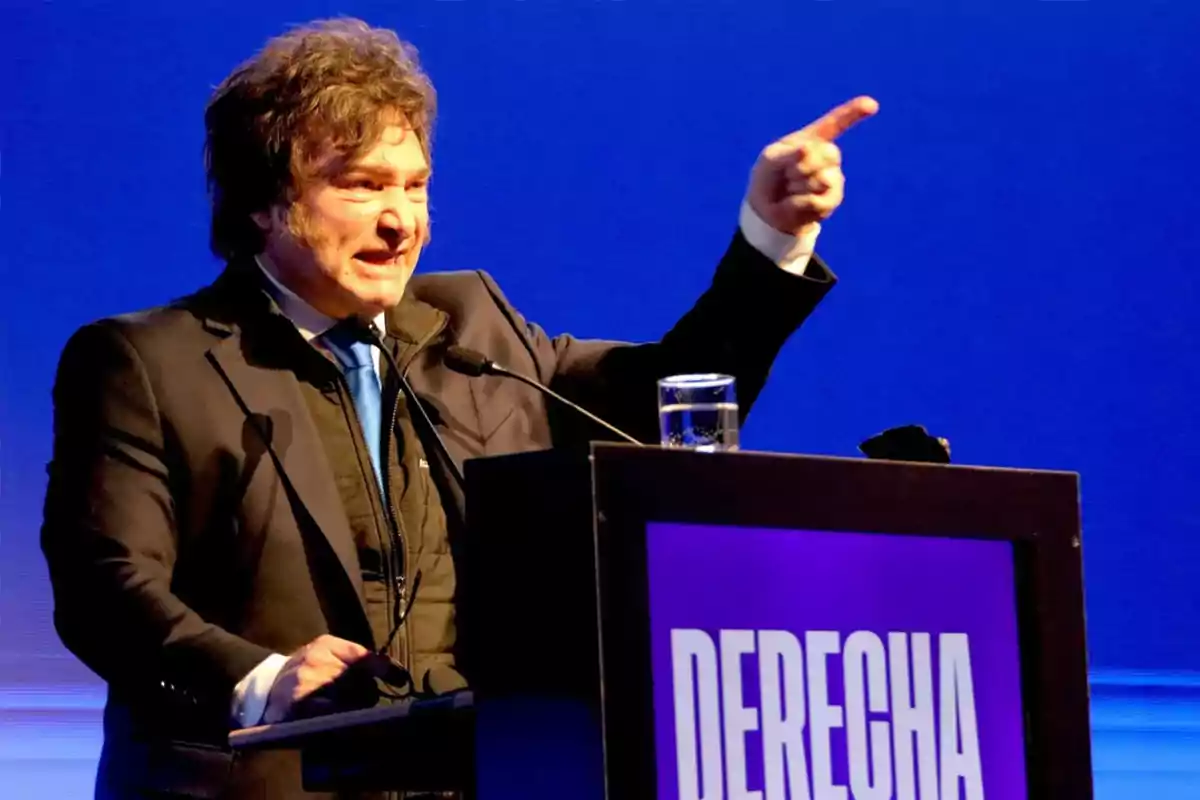 Hombre de cabello despeinado hablando en un podio con la palabra DERECHA y señalando con el dedo hacia el frente sobre un fondo azul Hombre de cabello despeinado hablando en un podio con la palabra DERECHA y señalando con el dedo hacia el frente sobre un fondo azul