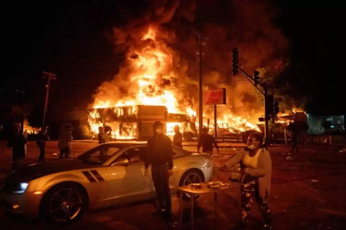 Personas en la calle durante la noche frente a un edificio envuelto en llamas con humo denso y un automóvil deportivo estacionado en primer plano