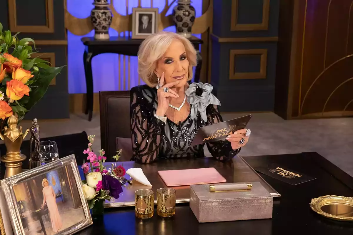 An elegantly dressed elderly woman is sitting at a table decorated with flowers, candles, and a framed photograph, holding a card with the text ML Mirtha in a luxurious television studio.