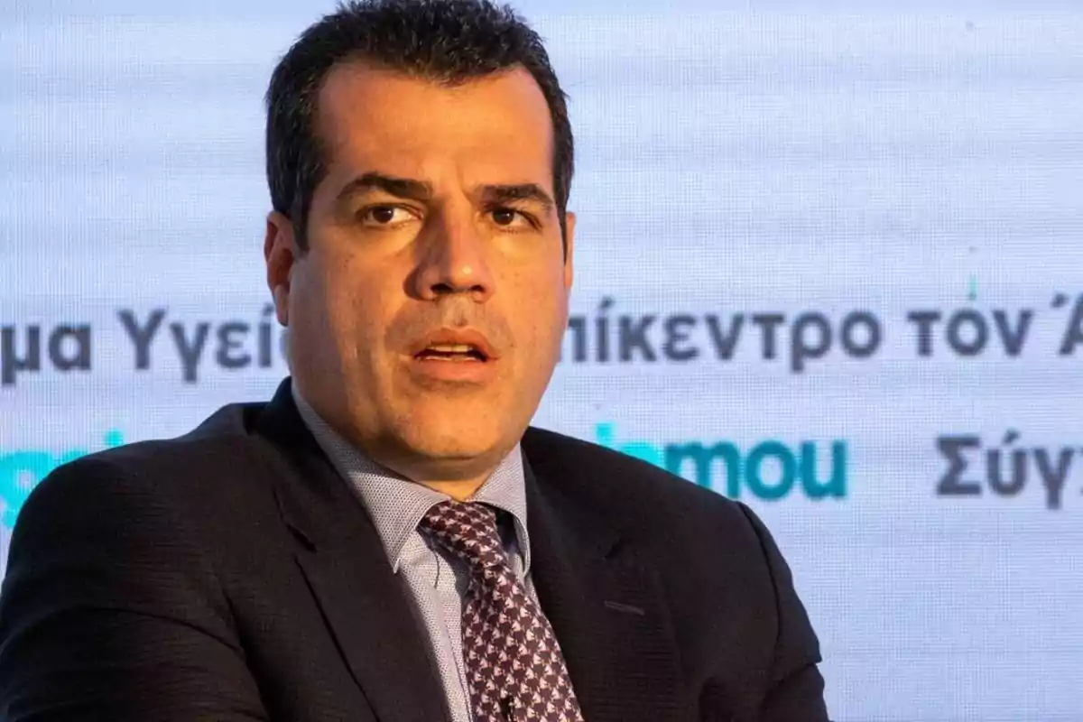 Hombre de cabello oscuro con traje y corbata sentado frente a una pantalla con texto en griego Hombre de cabello oscuro con traje y corbata sentado frente a una pantalla con texto en griego