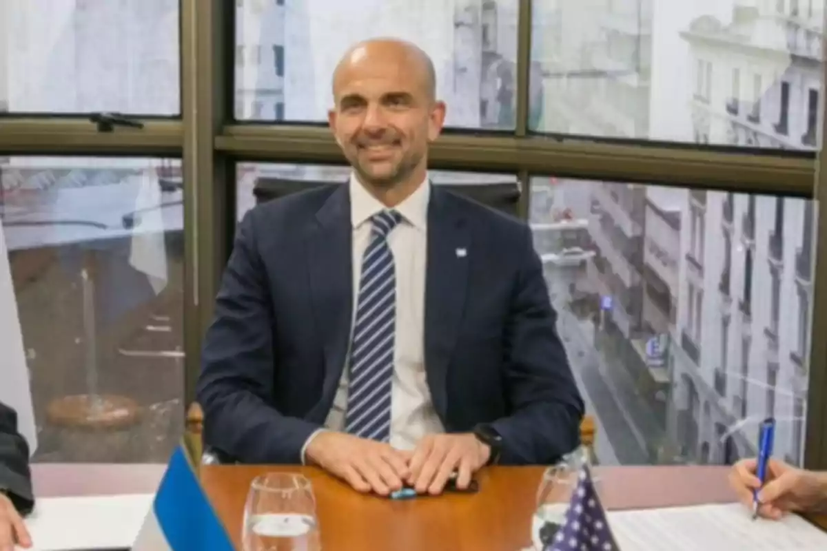 Bald man in a suit and tie sitting at a conference table with flags and documents, in an office with large windows and a view of the city Bald man in a suit and tie sitting at a conference table with flags and documents, in an office with large windows and a view of the city