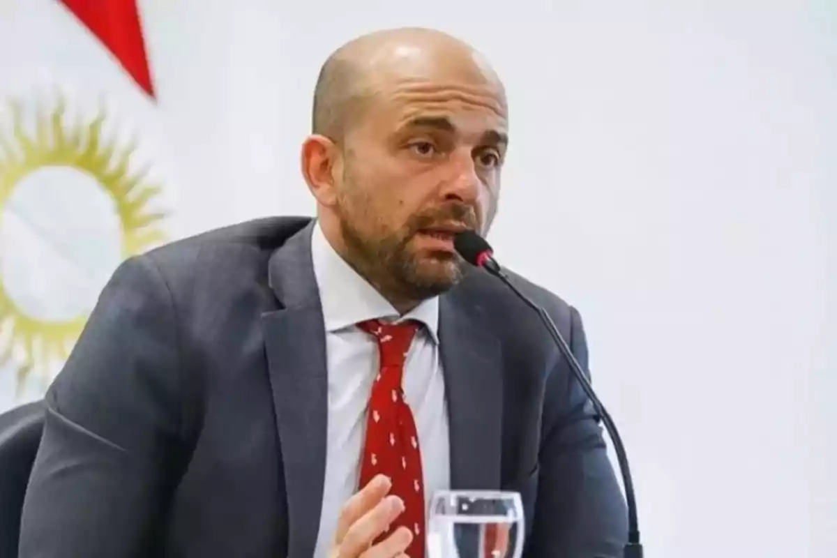 Bald man with a beard speaking into a microphone during a conference, wearing a gray suit and red tie, with a flag visible in the background.