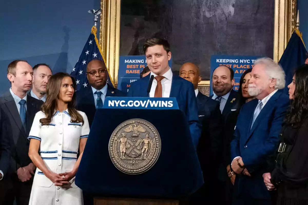 Un grupo de personas vestidas formalmente rodea a un hombre que habla en un podio con el escudo de la ciudad de Nueva York y un letrero que dice NYC.GOV/WINS, al fondo hay banderas y carteles informativos. Un grupo de personas vestidas formalmente rodea a un hombre que habla en un podio con el escudo de la ciudad de Nueva York y un letrero que dice NYC.GOV/WINS, al fondo hay banderas y carteles informativos.