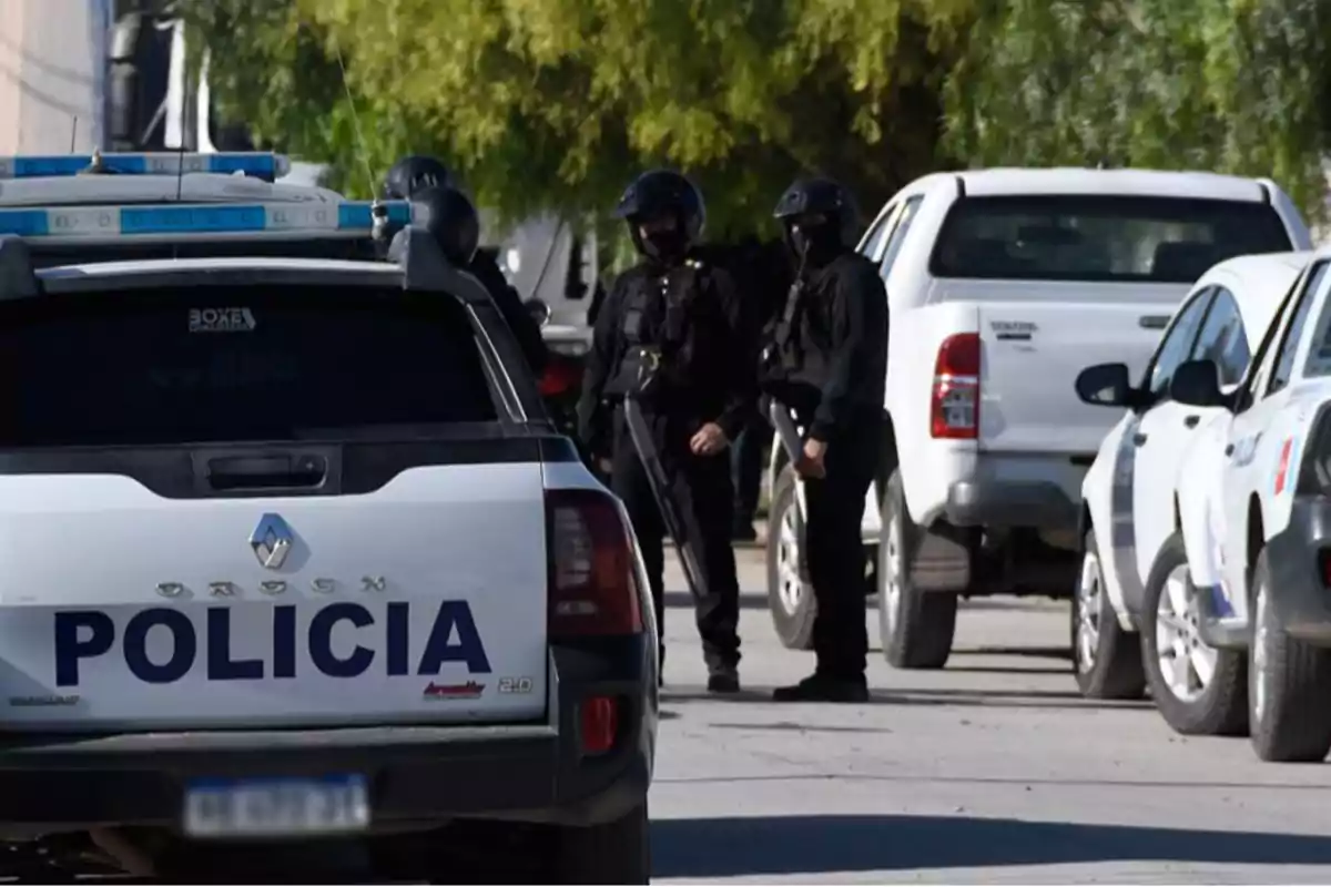 Police patrol car parked on a street next to several vehicles while three officers in uniform and tactical gear stand in the background.