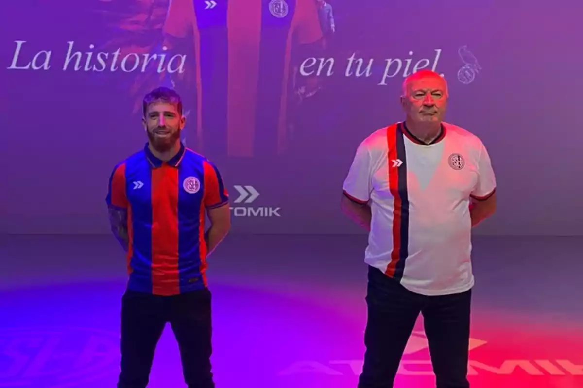 Dos hombres posan usando camisetas de fútbol del club San Luis, uno con la camiseta titular azul y roja y el otro con la camiseta alternativa blanca con una franja azul y roja, sobre un fondo violeta con la frase La historia en tu piel. Dos hombres posan usando camisetas de fútbol del club San Luis, uno con la camiseta titular azul y roja y el otro con la camiseta alternativa blanca con una franja azul y roja, sobre un fondo violeta con la frase La historia en tu piel.