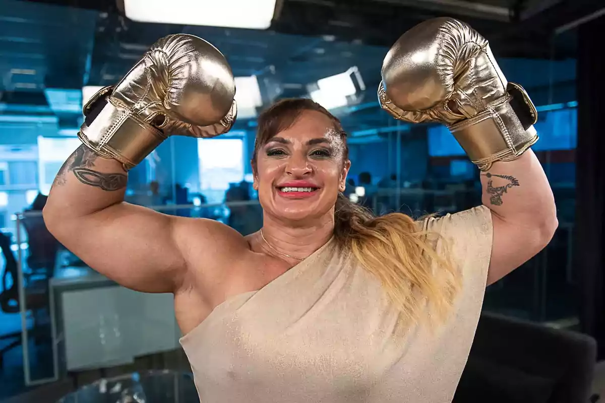 Mujer sonriente con guantes de box dorados levantando ambos brazos en señal de victoria dentro de una oficina moderna Mujer sonriente con guantes de box dorados levantando ambos brazos en señal de victoria dentro de una oficina moderna