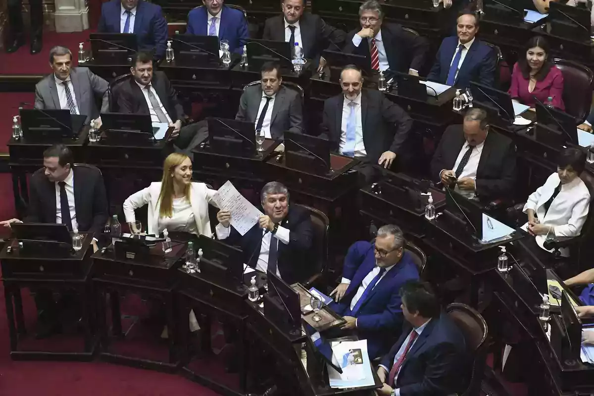 Several legislators seated at their desks during a parliamentary session, some conversing and others reviewing documents Several legislators seated at their desks during a parliamentary session, some conversing and others reviewing documents