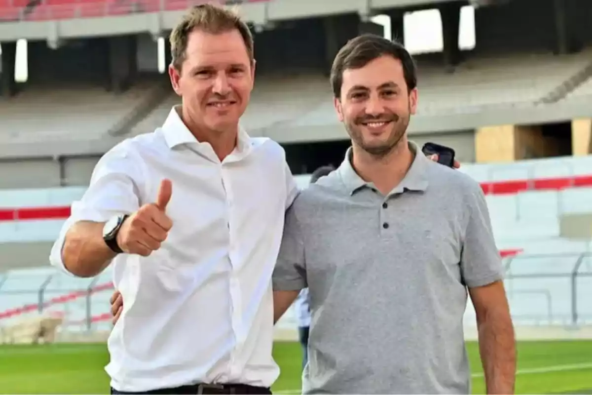 Two men smile and pose together in a stadium, one of them giving a thumbs-up gesture.