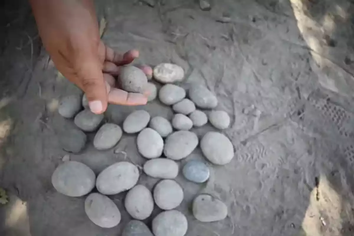 Mano sosteniendo una piedra sobre un grupo de piedras redondas acomodadas en el suelo de tierra Mano sosteniendo una piedra sobre un grupo de piedras redondas acomodadas en el suelo de tierra