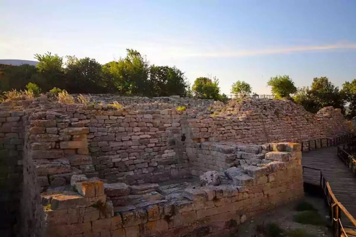 Ruinas de una antigua muralla de piedra rodeada de árboles y un camino de madera bajo un cielo despejado Ruinas de una antigua muralla de piedra rodeada de árboles y un camino de madera bajo un cielo despejado
