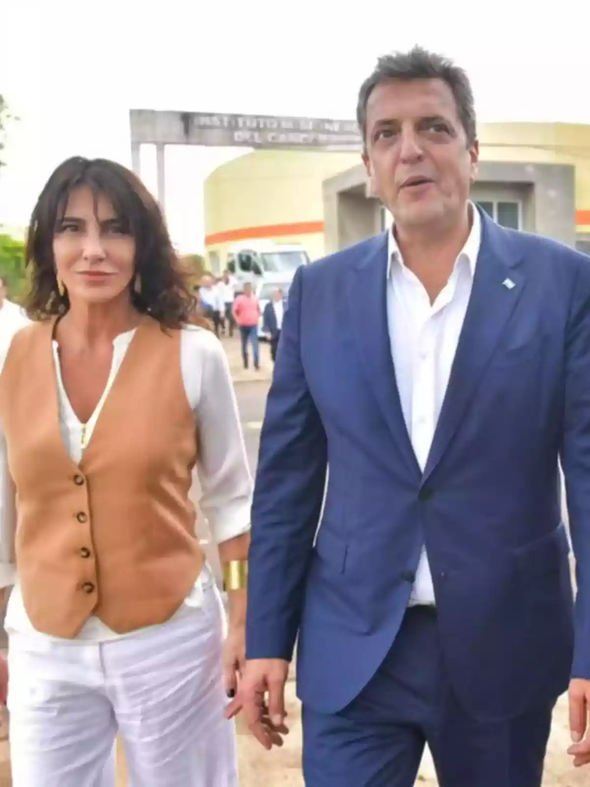 Una mujer con chaleco marrón y pantalón blanco camina junto a un hombre con traje azul en un entorno al aire libre con personas y un edificio al fondo Una mujer con chaleco marrón y pantalón blanco camina junto a un hombre con traje azul en un entorno al aire libre con personas y un edificio al fondo