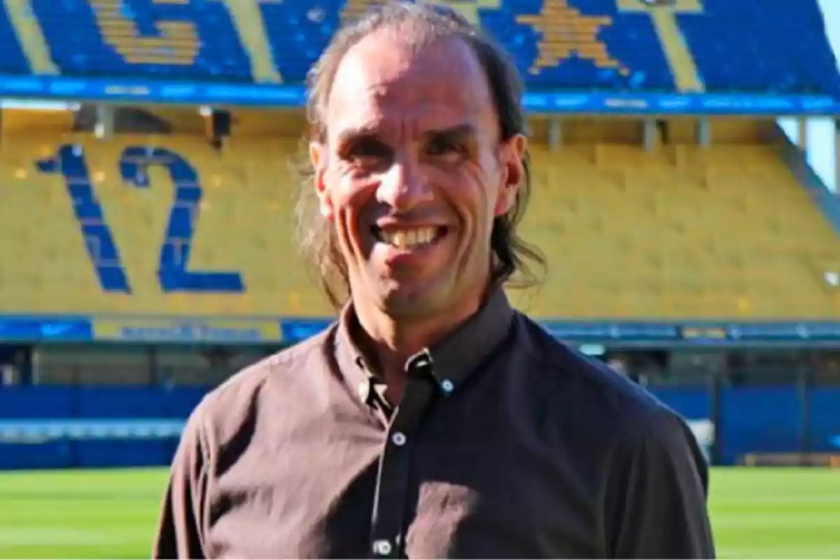 Hombre sonriente de cabello largo y camisa oscura posando en un estadio de fútbol con gradas amarillas y azules de fondo Hombre sonriente de cabello largo y camisa oscura posando en un estadio de fútbol con gradas amarillas y azules de fondo