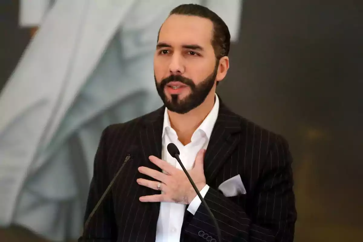 Hombre de barba y cabello recogido usando traje oscuro con rayas y camisa blanca hablando frente a dos micrófonos y con una mano en el pecho