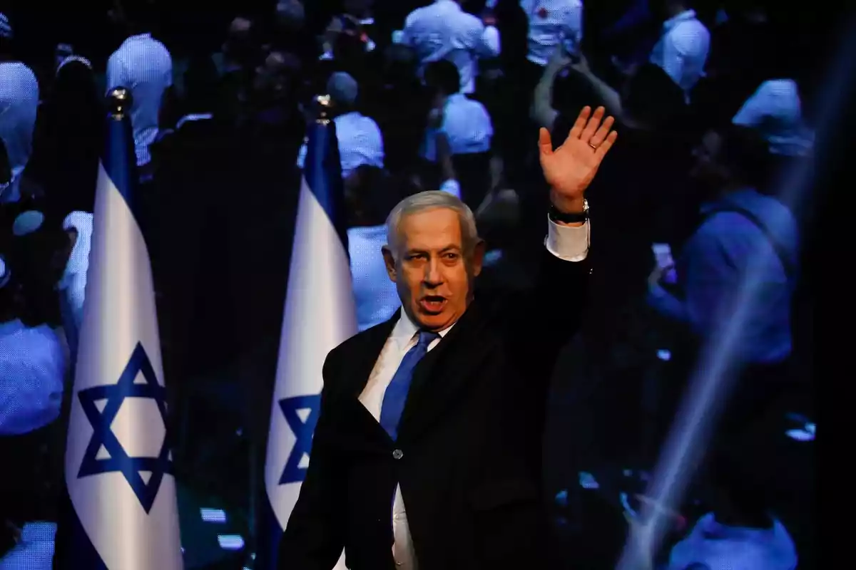 Hombre de cabello canoso con traje oscuro y corbata azul levantando la mano frente a dos banderas de Israel y un fondo de personas desenfocadas