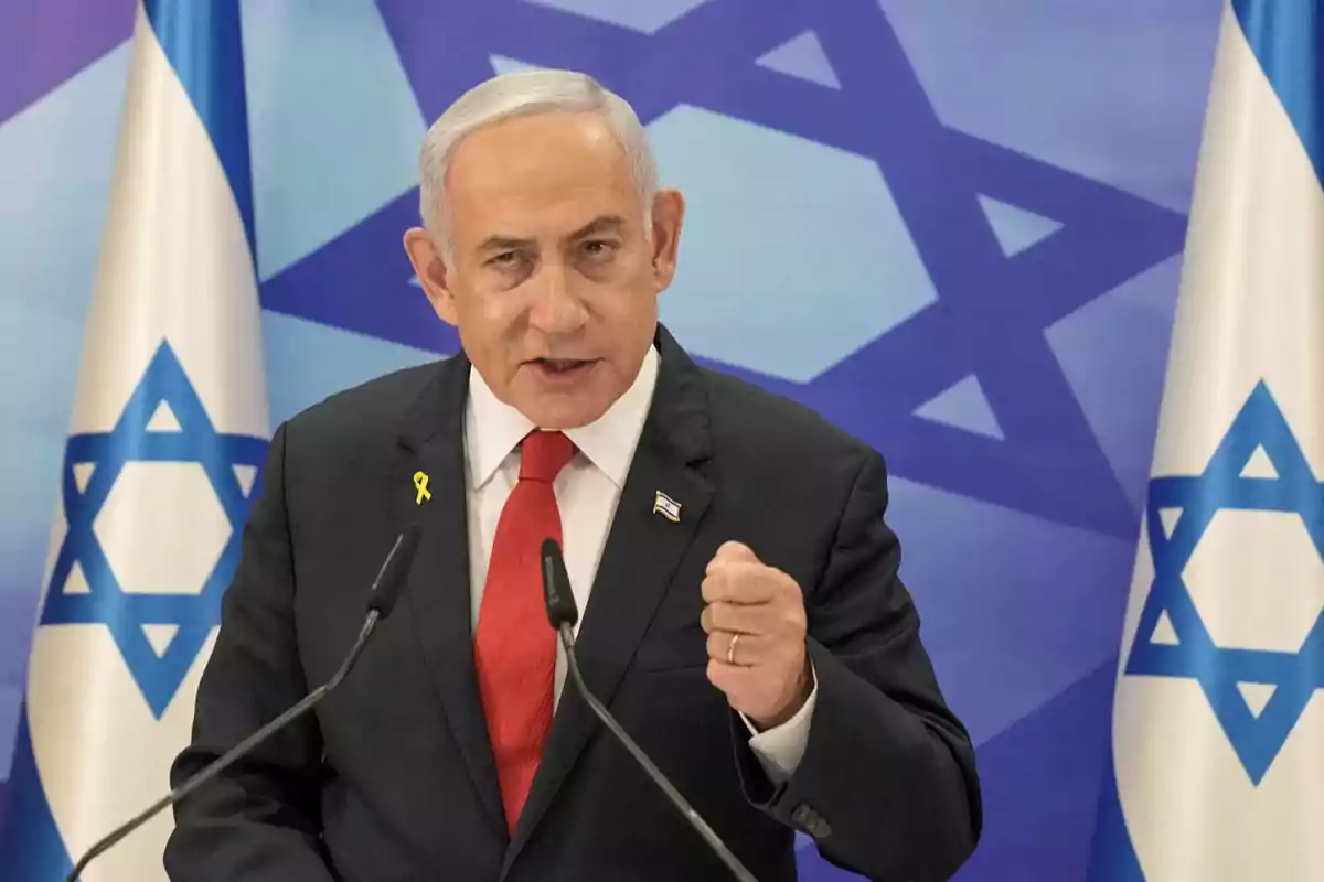 Gray-haired man in a dark suit and red tie speaking in front of microphones with Israeli flags in the background Gray-haired man in a dark suit and red tie speaking in front of microphones with Israeli flags in the background