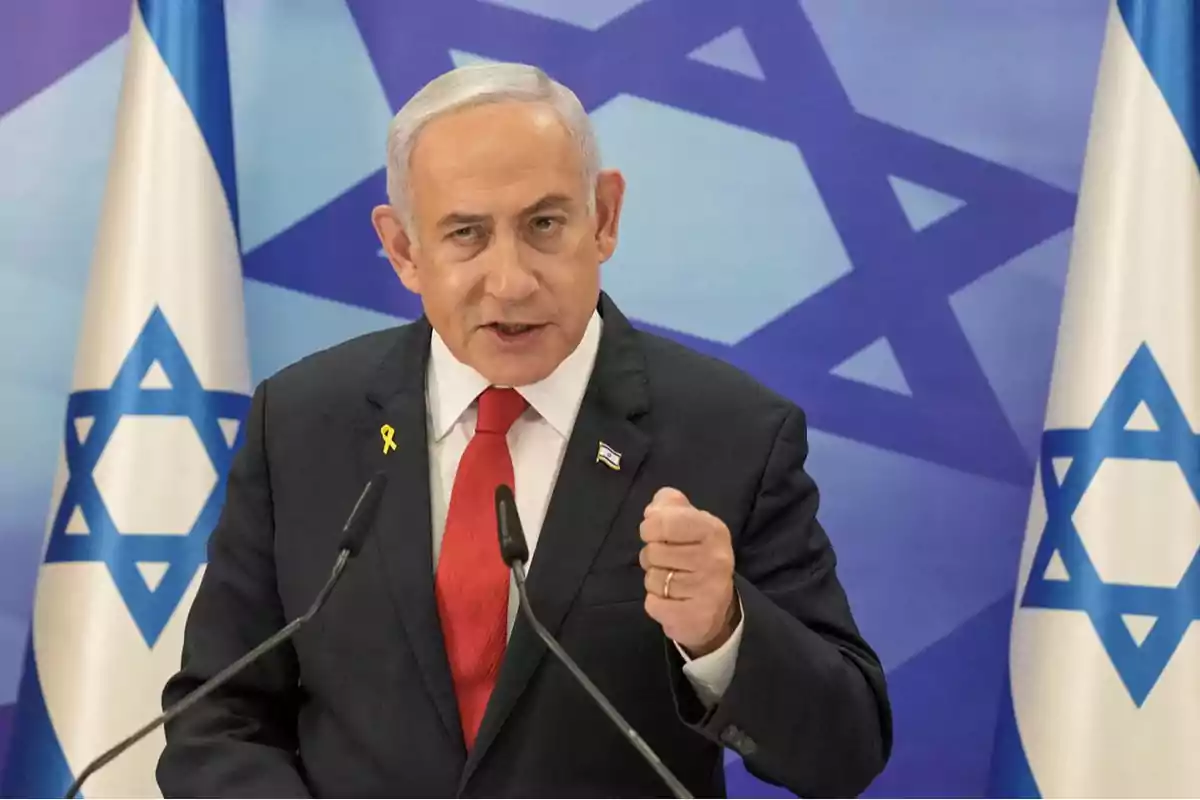 Hombre de cabello canoso con traje oscuro y corbata roja hablando frente a micrófonos con banderas de Israel de fondo