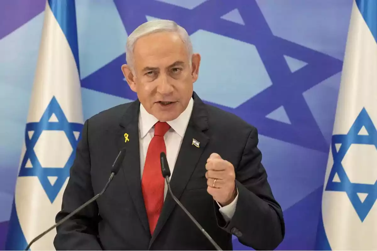 Hombre de cabello canoso con traje oscuro y corbata roja hablando frente a micrófonos con banderas de Israel de fondo Hombre de cabello canoso con traje oscuro y corbata roja hablando frente a micrófonos con banderas de Israel de fondo