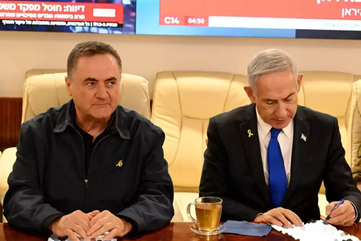 Two men are sitting in light-colored armchairs; one of them is writing on some papers while the other watches. Both are wearing a yellow ribbon on their lapel, and there is a cup of tea on the table.