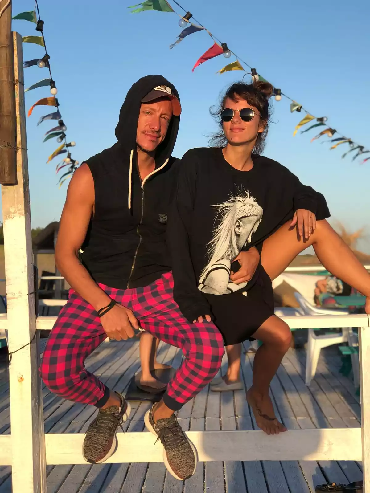 Dos personas sentadas en una estructura de madera al aire libre con banderines de colores y cielo despejado de fondo, ambos visten ropa casual y gafas de sol Dos personas sentadas en una estructura de madera al aire libre con banderines de colores y cielo despejado de fondo, ambos visten ropa casual y gafas de sol