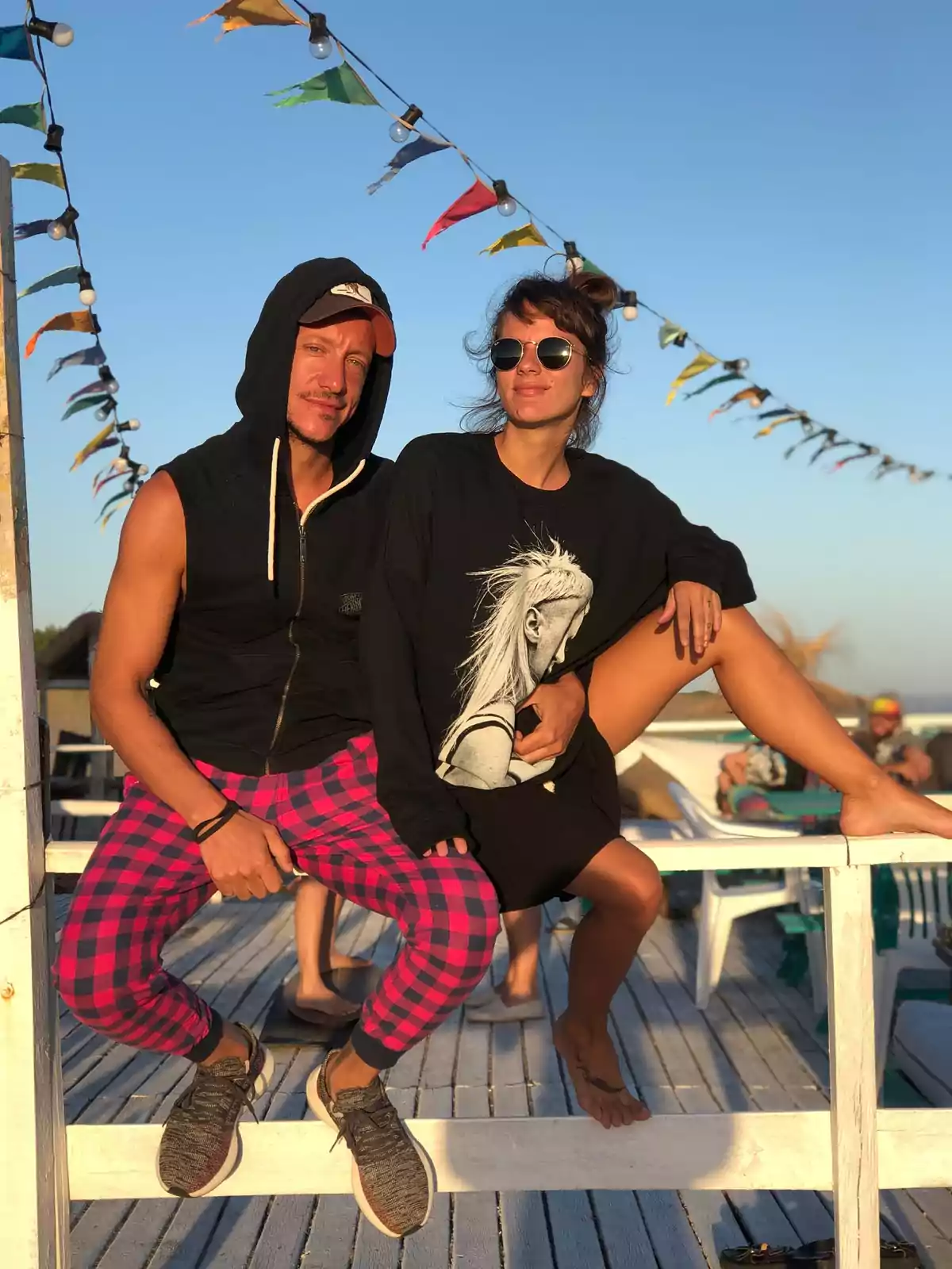 Dos personas posan sentadas sobre una baranda de madera en un ambiente al aire libre con banderines de colores y cielo despejado, ambos lucen ropa casual y gafas de sol Dos personas posan sentadas sobre una baranda de madera en un ambiente al aire libre con banderines de colores y cielo despejado, ambos lucen ropa casual y gafas de sol