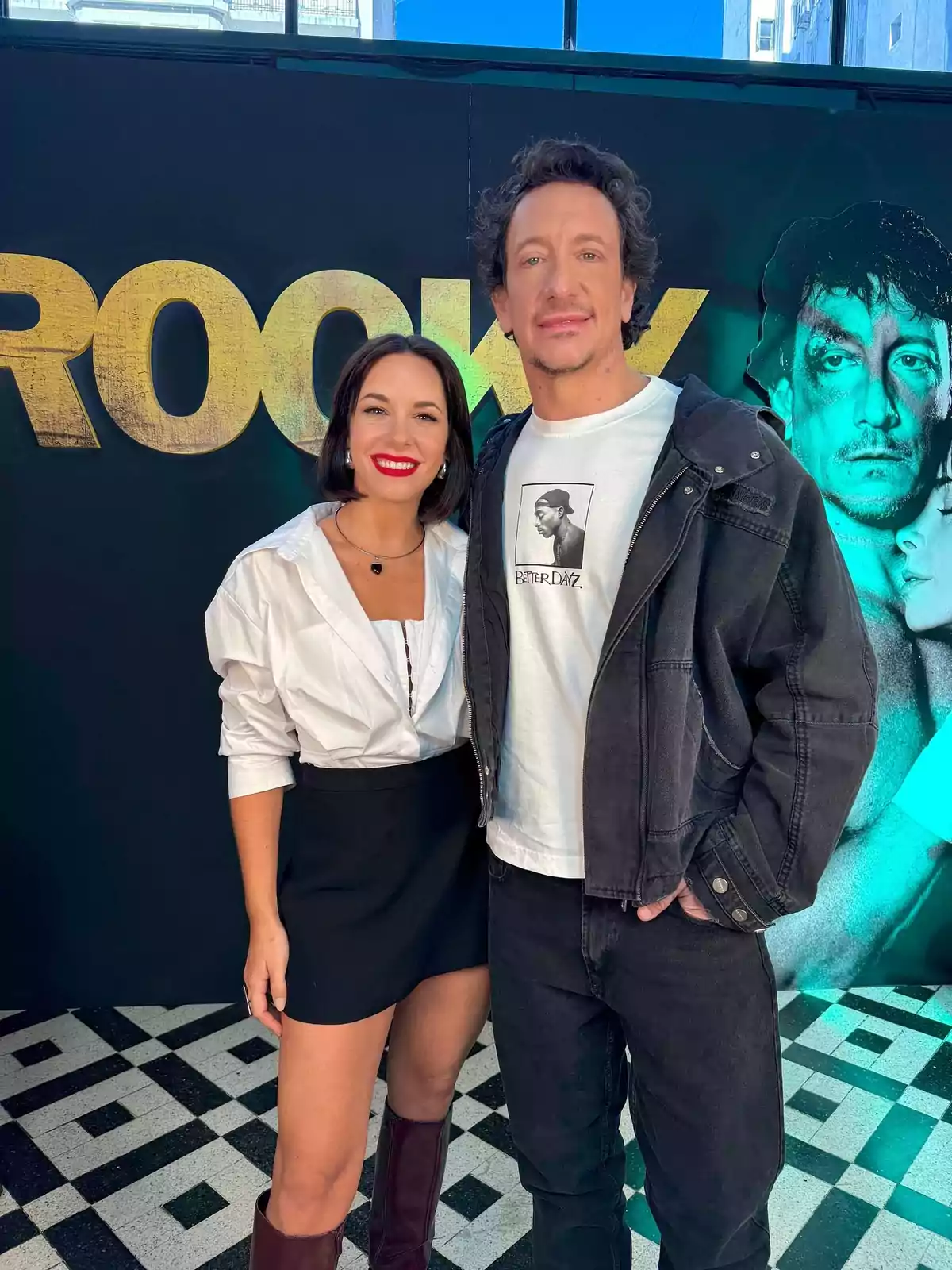 Two people pose smiling in front of a promotional backdrop for the movie Rocky, both dressed casually and elegantly, on a black and white mosaic floor. Two people pose smiling in front of a promotional backdrop for the movie Rocky, both dressed casually and elegantly, on a black and white mosaic floor.