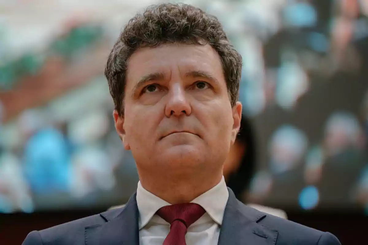 Hombre de cabello rizado y traje formal con corbata roja mirando hacia arriba con expresión seria y fondo desenfocado