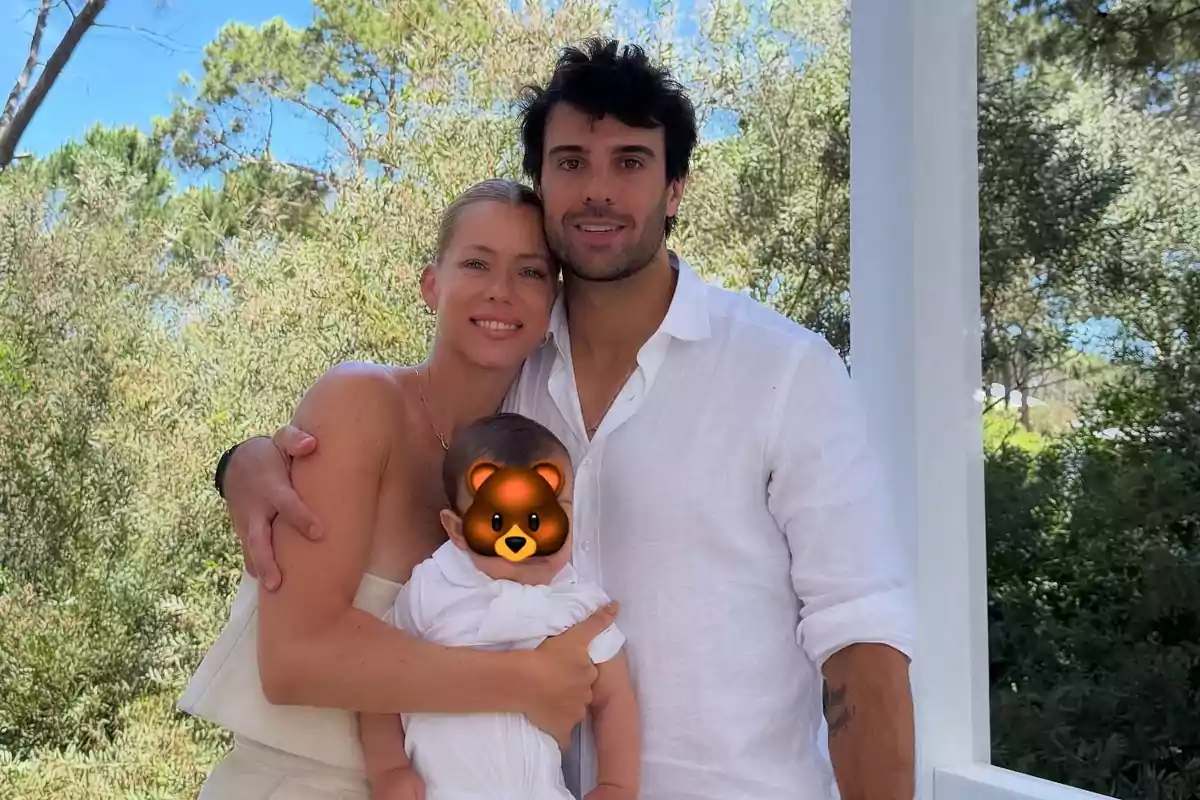 Pareja abrazando a un niño pequeño cuyo rostro está cubierto con un emoji de oso, todos vestidos de blanco y posando al aire libre rodeados de vegetación