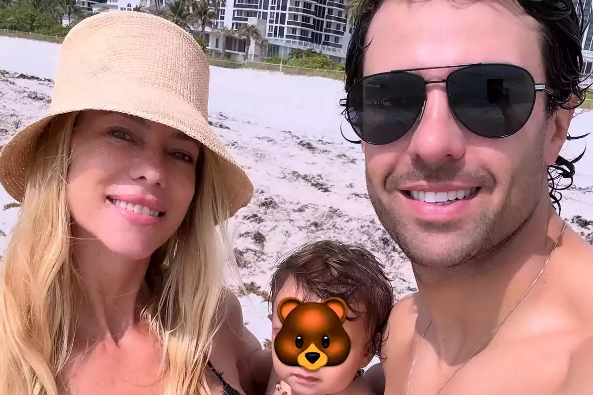 Familia en la playa sonriendo mientras disfrutan de un día soleado, con edificios al fondo y el rostro del niño cubierto con un emoji de oso Familia en la playa sonriendo mientras disfrutan de un día soleado, con edificios al fondo y el rostro del niño cubierto con un emoji de oso