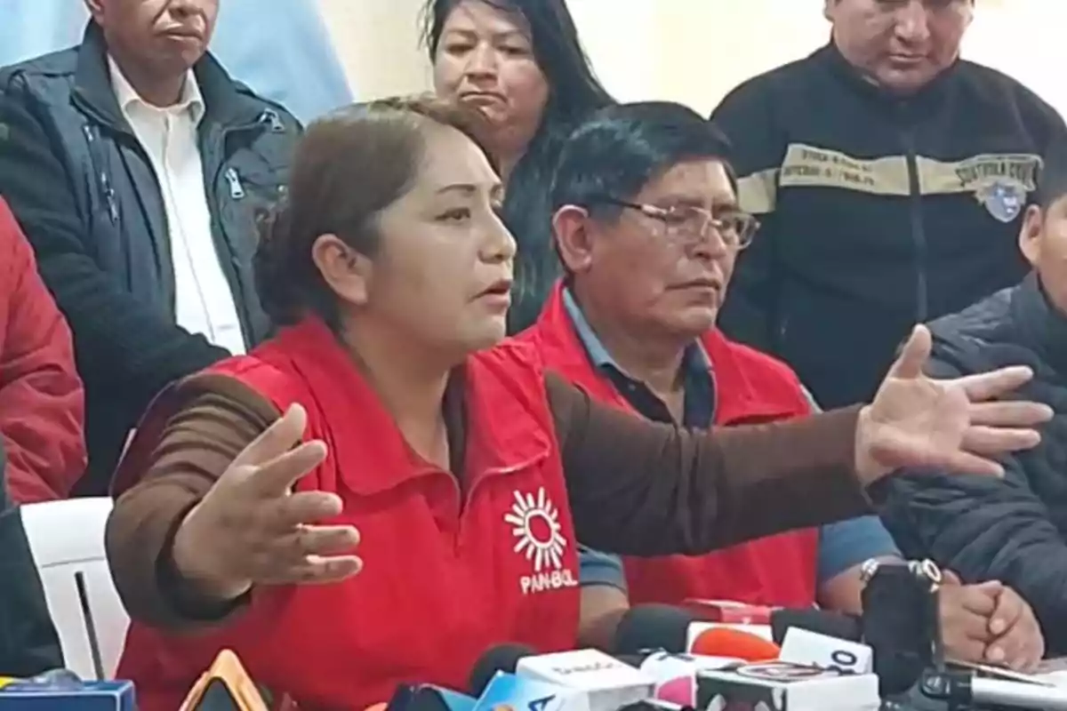 Mujer con chaleco rojo hablando en una conferencia de prensa rodeada de varias personas y micrófonos sobre la mesa
