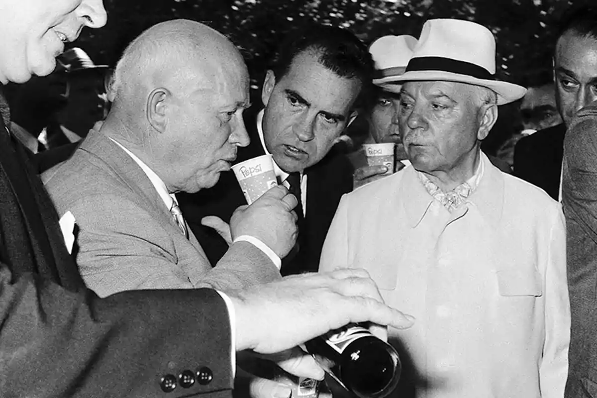 Several men dressed in suits and hats are talking while one of them drinks from a glass with the Pepsi logo in a black-and-white scene. Several men dressed in suits and hats are talking while one of them drinks from a glass with the Pepsi logo in a black-and-white scene.