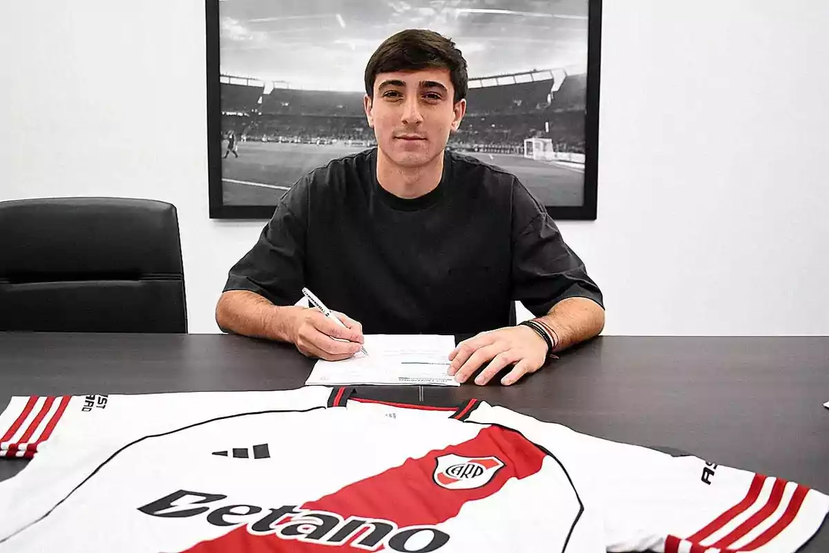 Joven firmando un contrato en una oficina con una camiseta de River Plate sobre la mesa
