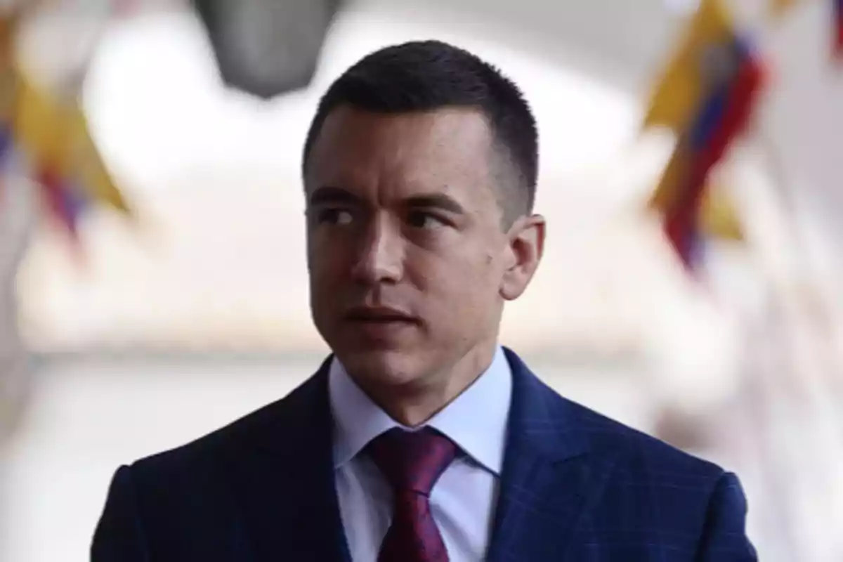Hombre de cabello corto y oscuro con traje azul y corbata roja mirando hacia un lado en un entorno formal con banderas desenfocadas al fondo