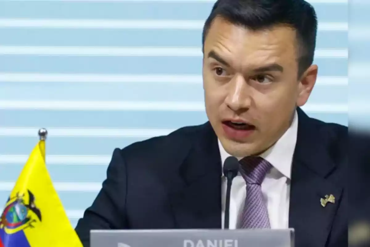 Hombre de traje oscuro y corbata lila hablando frente a un micrófono con una bandera de Ecuador a su lado