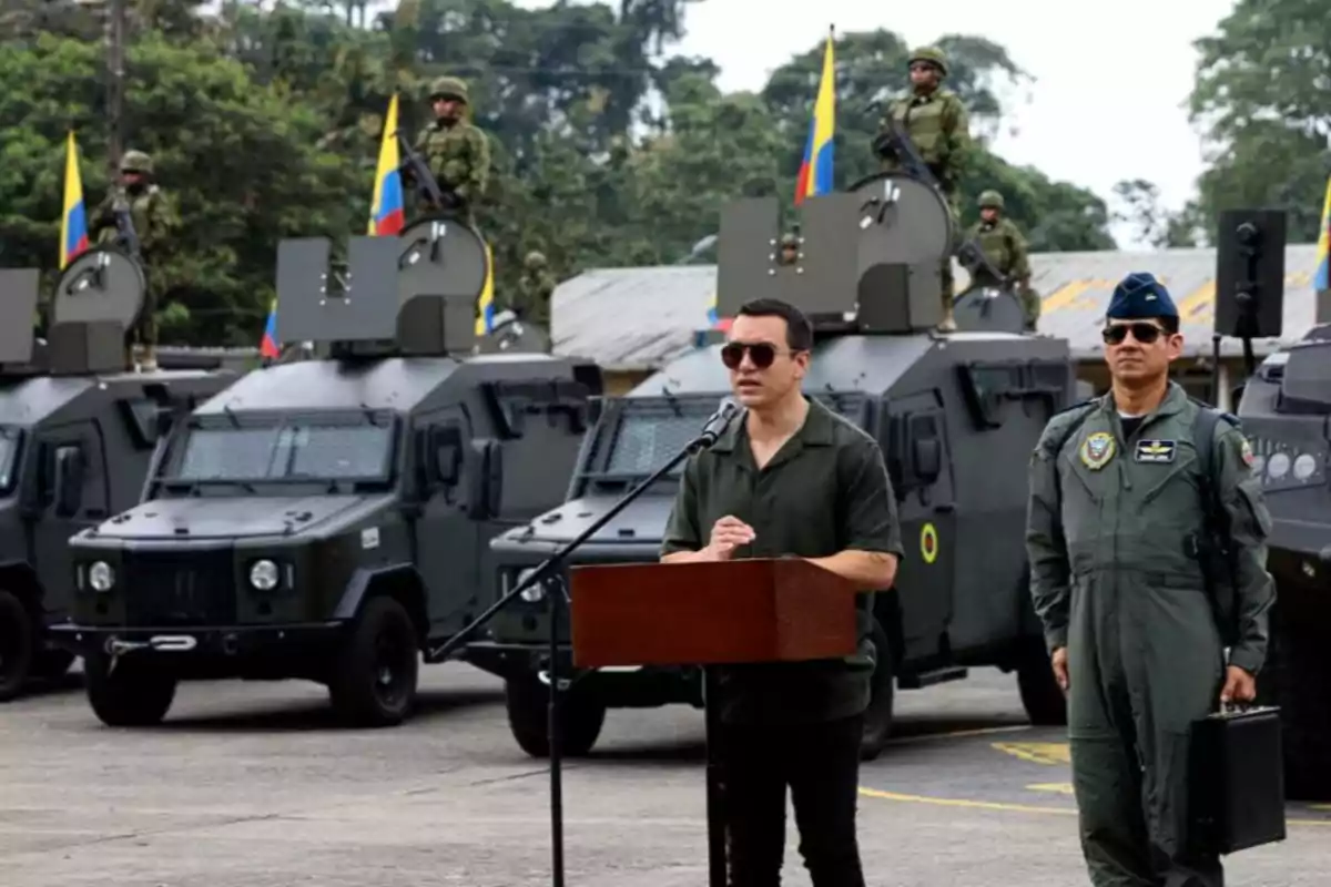 A man wearing dark glasses gives a speech in front of armored military vehicles while armed soldiers and Colombian flags are on the vehicles and an air force officer stands by his side.