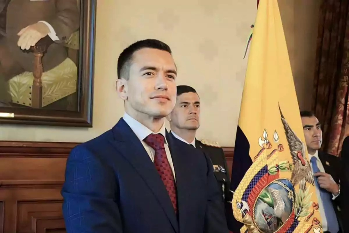 Man in a blue suit and red tie standing next to an Ecuadorian flag in a formal setting with other people in the background Man in a blue suit and red tie standing next to an Ecuadorian flag in a formal setting with other people in the background