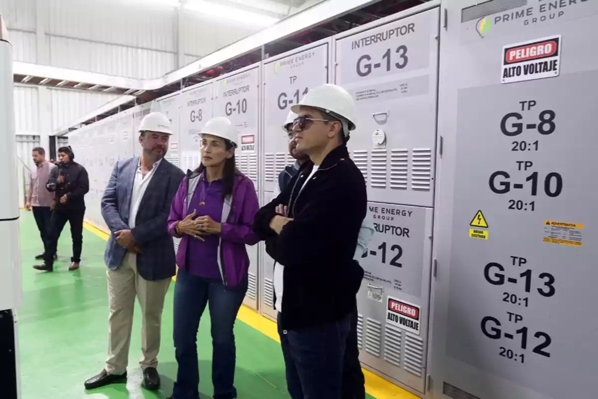Varias personas con casco de seguridad recorren una planta industrial con paneles eléctricos y señales de alto voltaje Varias personas con casco de seguridad recorren una planta industrial con paneles eléctricos y señales de alto voltaje