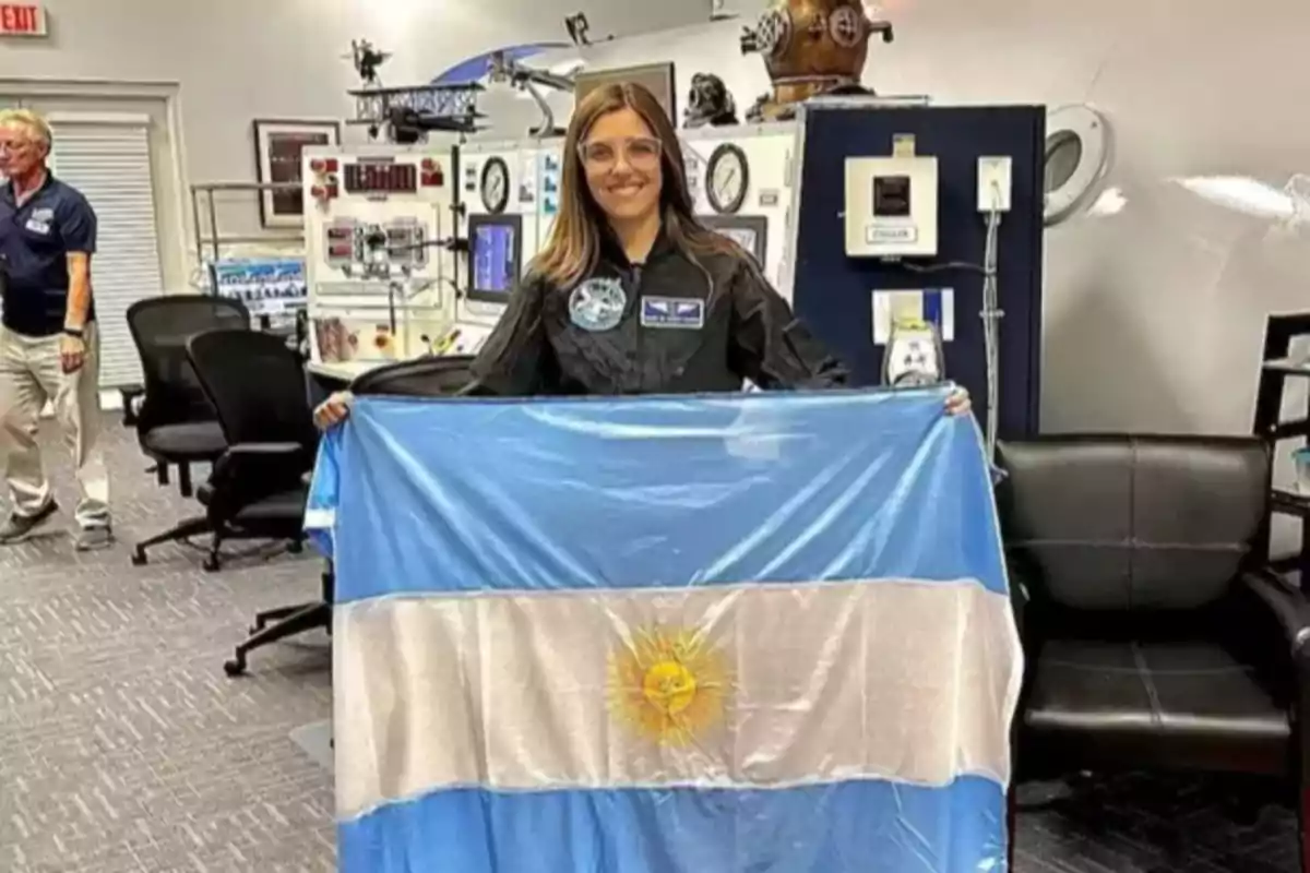 Mujer sonriente con gafas y traje negro sostiene una bandera de Argentina en una sala con equipos tecnológicos y muebles de oficina Mujer sonriente con gafas y traje negro sostiene una bandera de Argentina en una sala con equipos tecnológicos y muebles de oficina