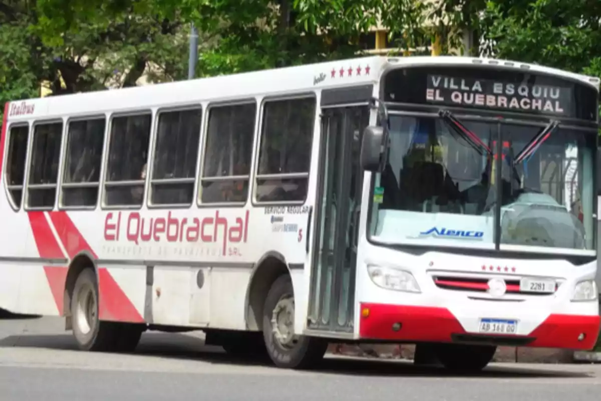 A white and red bus from the company El Quebrachal stopped on a street with trees in the background A white and red bus from the company El Quebrachal stopped on a street with trees in the background
