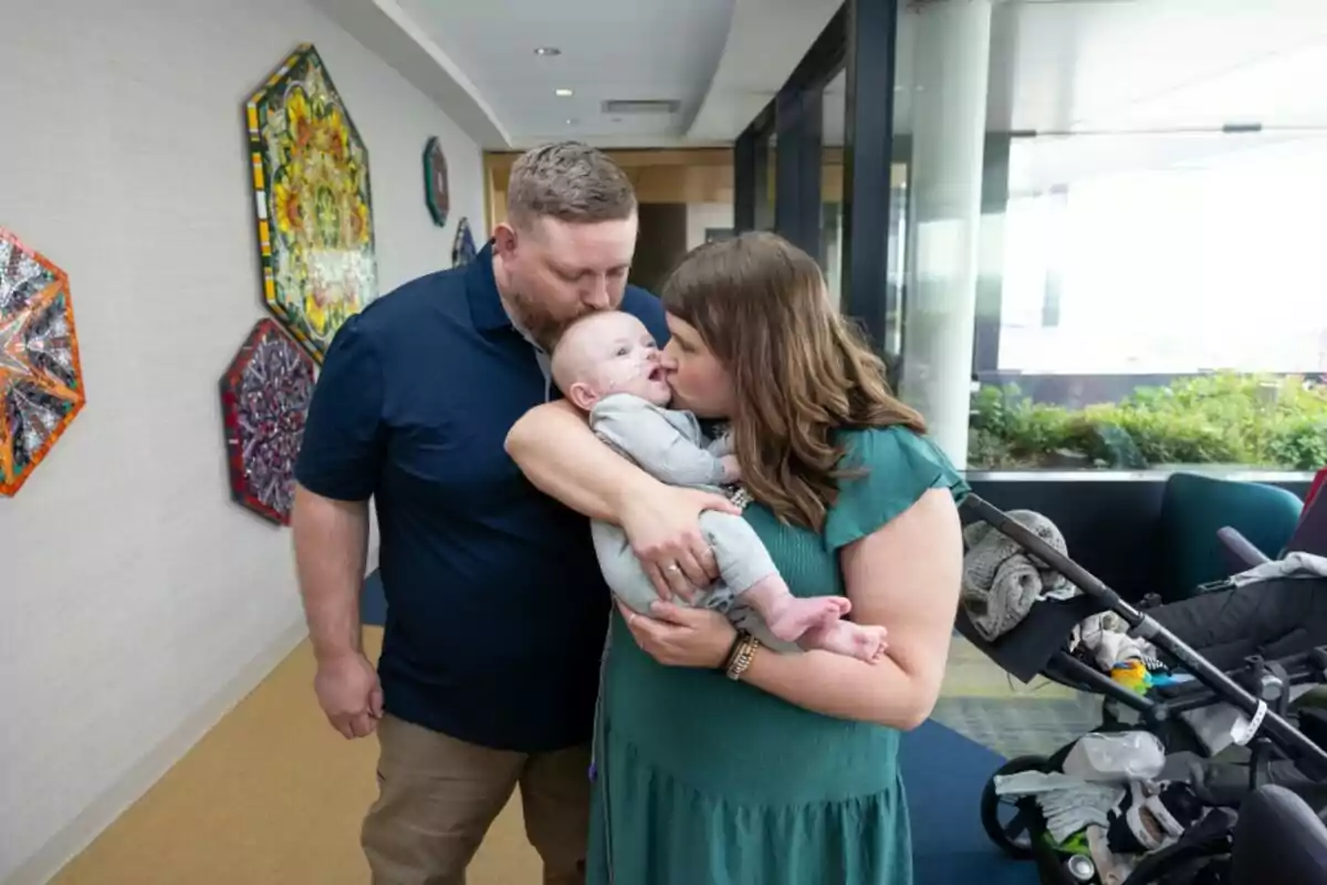 Una pareja sostiene y besa a un bebé en un pasillo decorado con coloridos mosaicos y un cochecito de bebé al fondo
