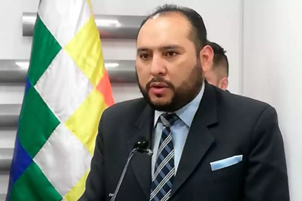 Hombre de barba y traje oscuro hablando frente a un micrófono con una bandera de fondo