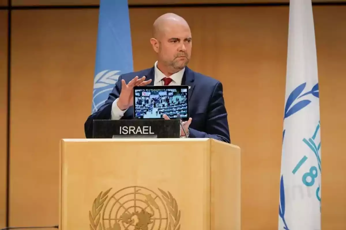Man in a suit behind a podium with the Israel sign holding a tablet in a room with UN flags.
