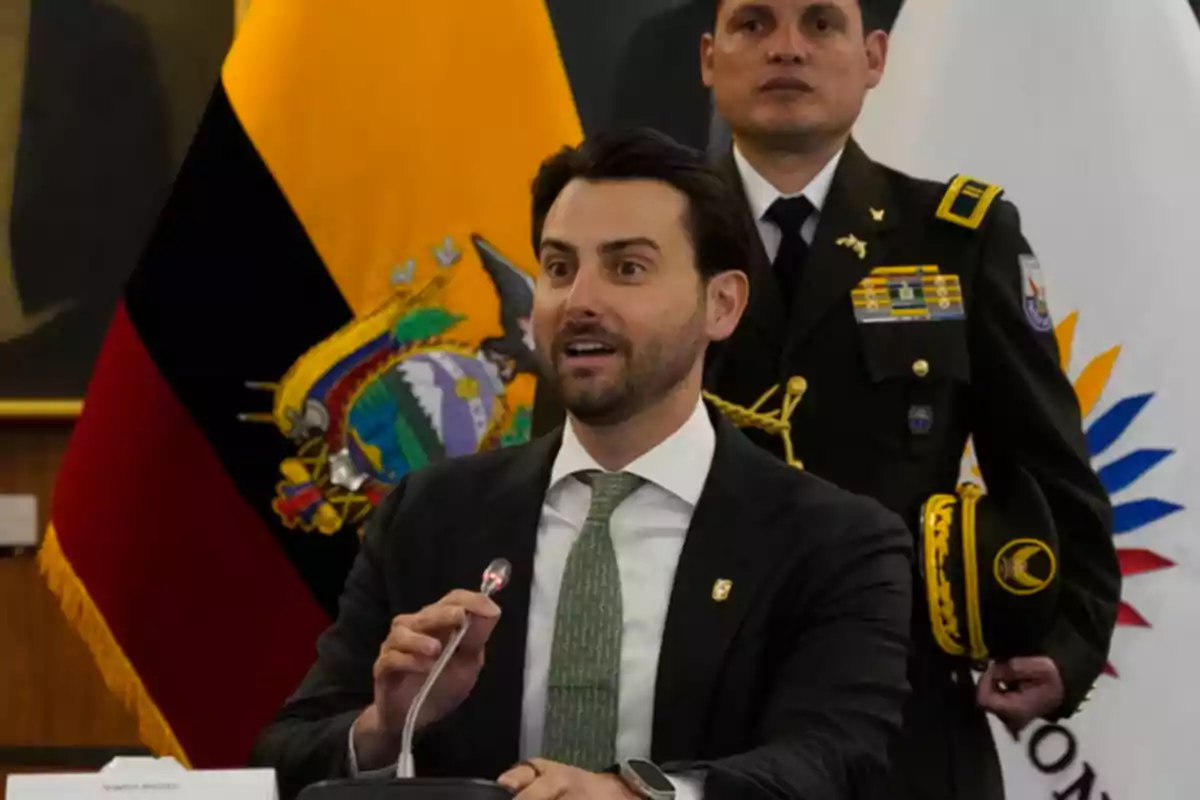Man in a suit speaking in front of a microphone with a military officer and Ecuadorian flags in the background