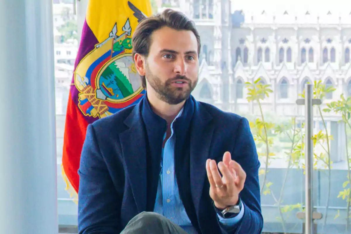 Hombre de barba y cabello oscuro vestido con saco azul sentado frente a una bandera de Ecuador y un edificio histórico al fondo mientras gesticula con la mano Hombre de barba y cabello oscuro vestido con saco azul sentado frente a una bandera de Ecuador y un edificio histórico al fondo mientras gesticula con la mano