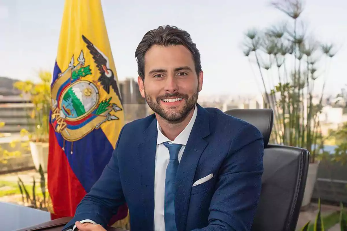 Hombre de traje azul sentado en una oficina con una bandera de Ecuador al fondo y plantas decorativas