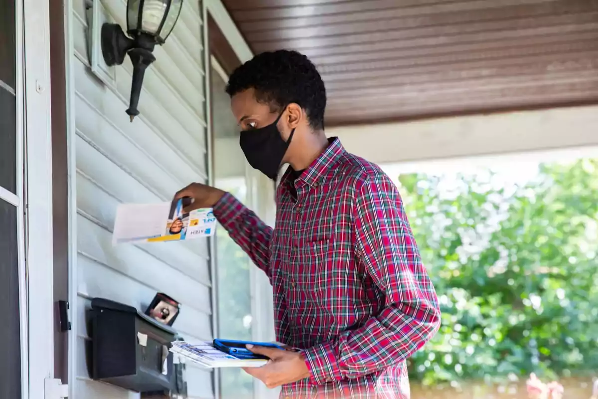Man wearing a mask and a plaid jersey dropping letters into a mailbox in front of a house Man wearing a mask and a plaid jersey dropping letters into a mailbox in front of a house