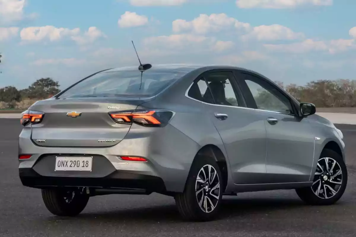 Automóvil sedán gris Chevrolet Onix visto desde la parte trasera estacionado en un espacio abierto con cielo azul y nubes de fondo Automóvil sedán gris Chevrolet Onix visto desde la parte trasera estacionado en un espacio abierto con cielo azul y nubes de fondo