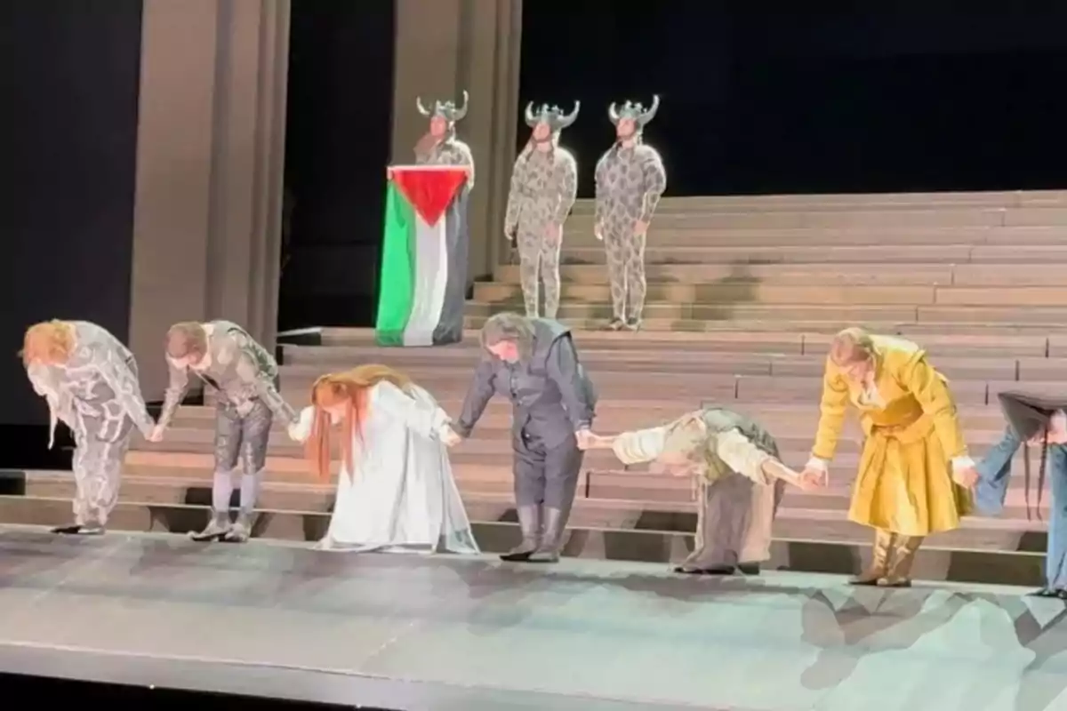 Un grupo de actores vestidos con trajes de época se inclina en señal de agradecimiento al público sobre un escenario mientras al fondo tres personas con disfraces de cuernos y una de ellas sostiene una bandera con los colores de Palestina