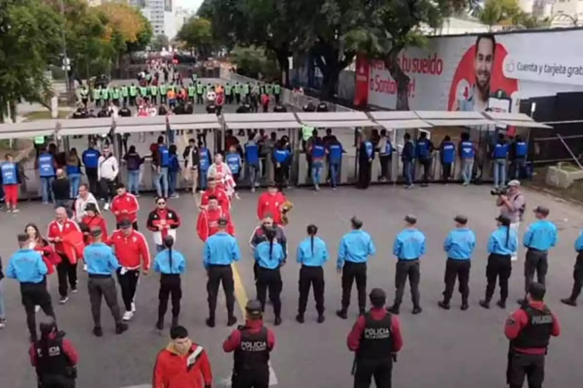 Aficionados vestidos de rojo pasan por un control de seguridad con presencia de policías y personal de seguridad en la entrada de un estadio Aficionados vestidos de rojo pasan por un control de seguridad con presencia de policías y personal de seguridad en la entrada de un estadio