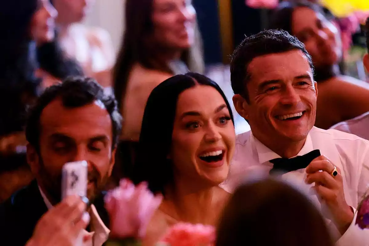 Personas sentadas en una mesa durante un evento elegante sonríen y se ven emocionadas mientras observan algo fuera de cámara