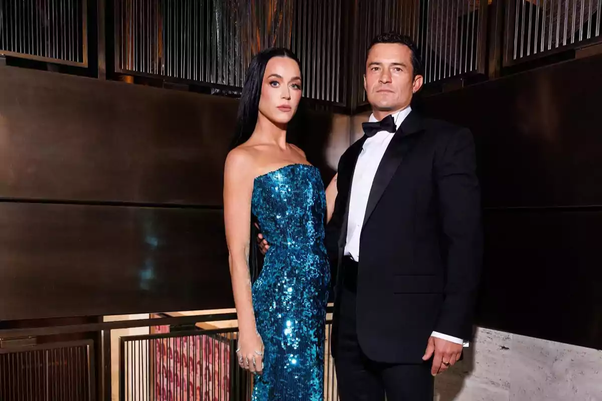 An elegantly dressed couple poses in front of a metallic wall; the woman is wearing a shiny blue dress and the man a black tuxedo.
